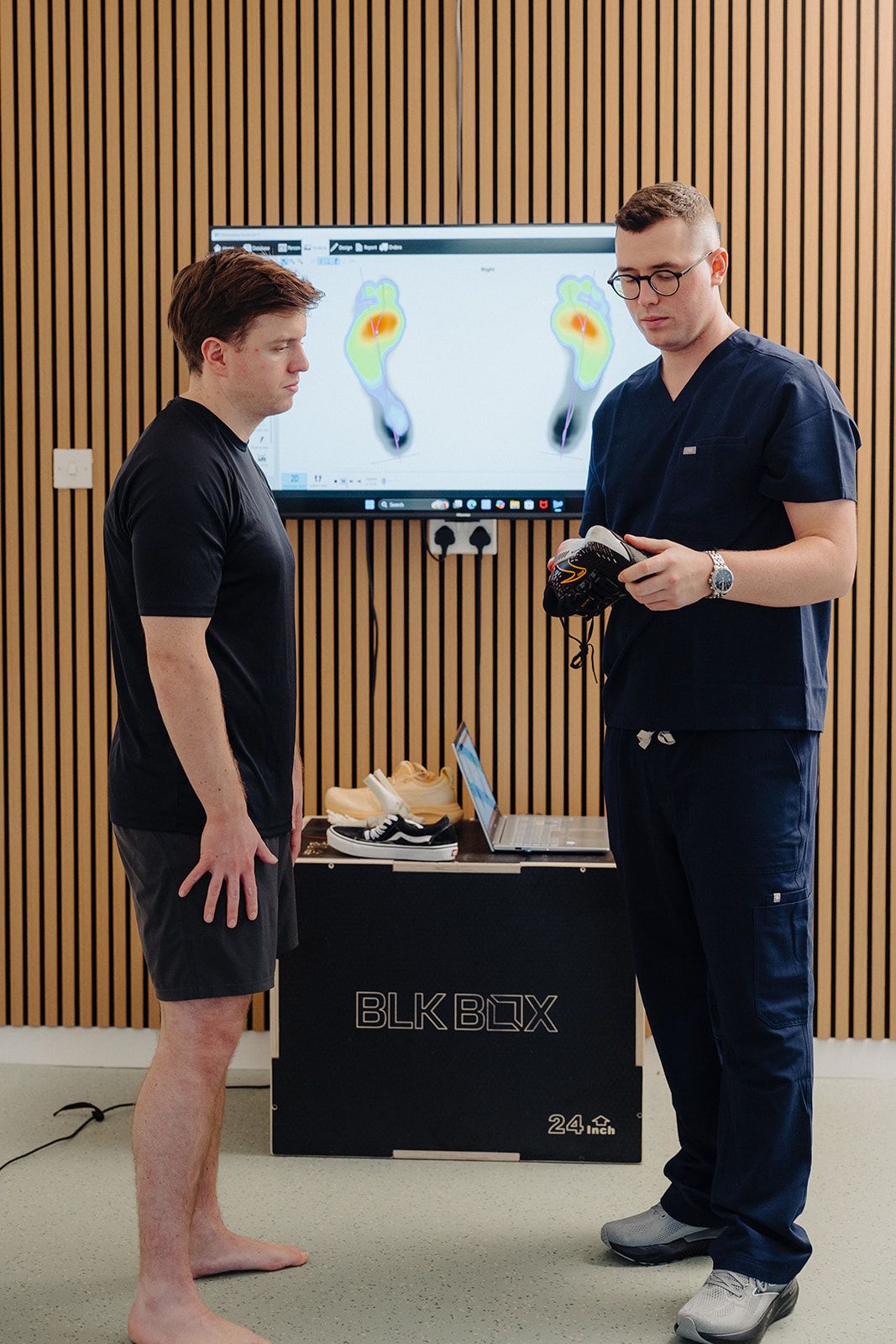 Podiatrist reviewing a patient's running shoe in front of Footscan pressure plate analysis data at Assure Health in Carryduff, Belfast
